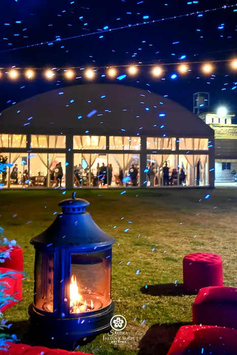 Chimenea de leña en el jardín con el salón de cristal iluminado al fondo